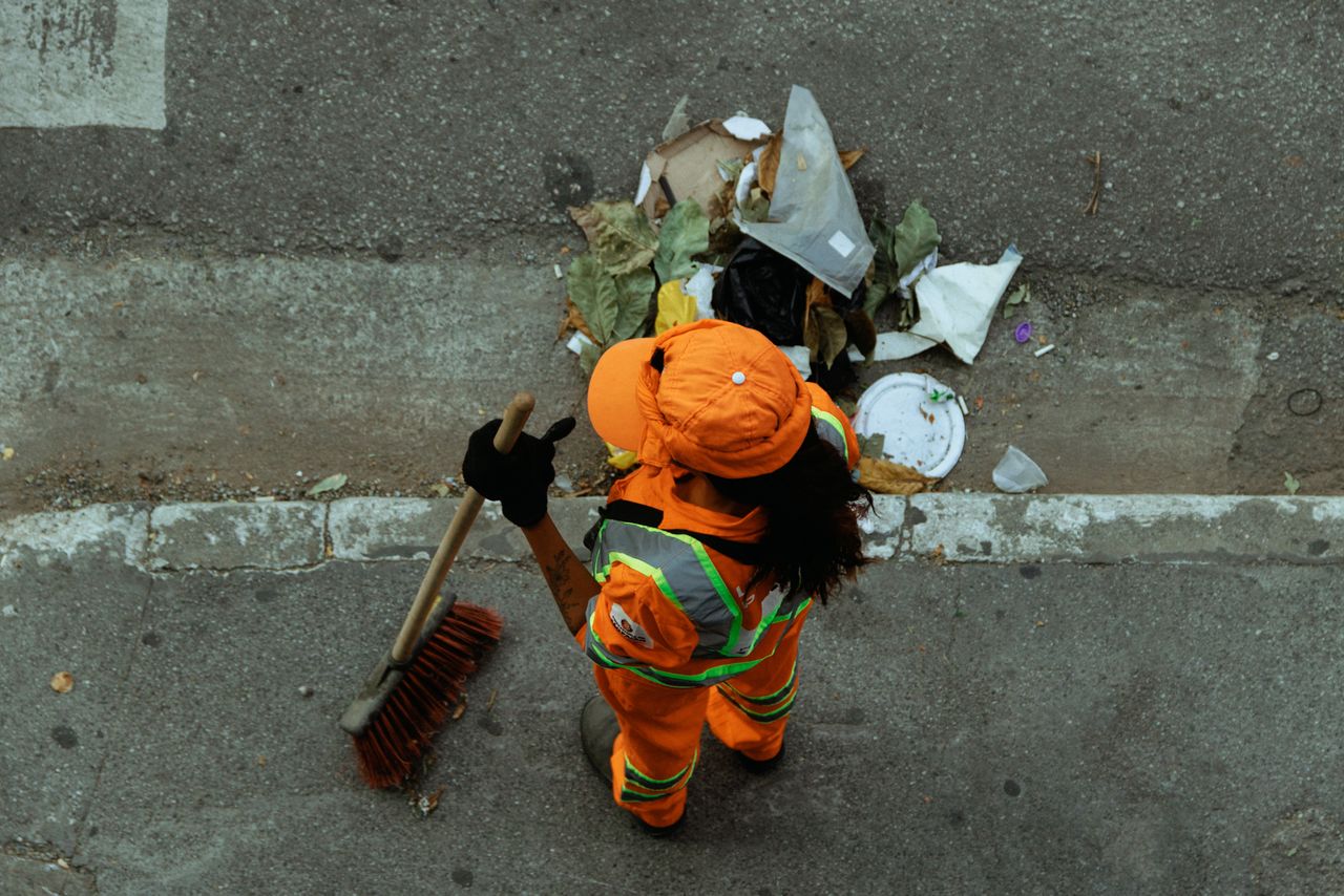 Home Top view of a street cleaner wearing orange uniform sweeping debris on a city sidewalk.
