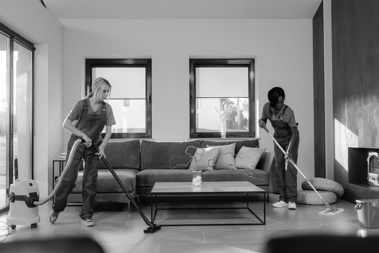 Two women in work clothes cleaning a modern living room using a vacuum and mop.